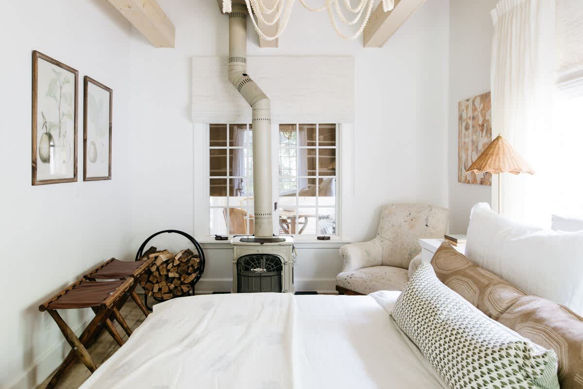 A bright bedroom features a white bed, a vintage-style wood-burning stove with a tall pipe, exposed wooden ceiling beams, and a window with a Roman shade, creating a cozy and rustic ambiance.