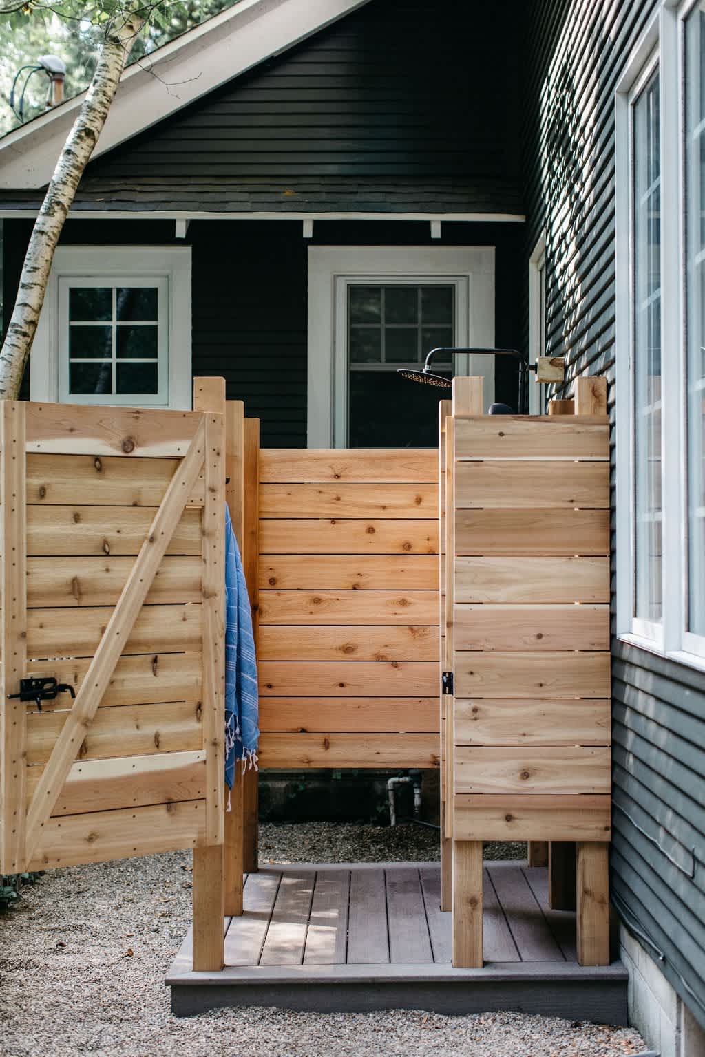 Rustic outdoor shower enclosure made of natural wood planks, featuring a black shower head and a blue towel hanging on the door, set against a dark-sided house.