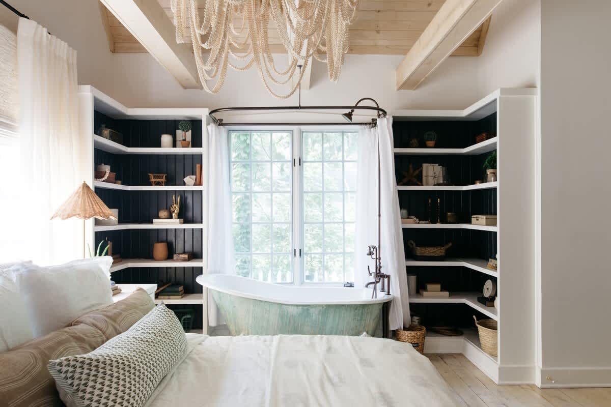 Serene master bedroom featuring a large bed with neutral bedding, exposed wooden ceiling beams, and a unique clawfoot tub with a distressed green finish positioned in front of a large window. Built-in shelves with dark backings flank the window, displaying decorative items.