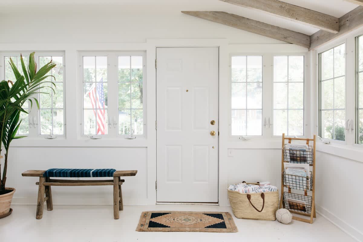 A bright, white entryway features multiple grid-paned windows, a white paneled door, a rustic wooden bench with a blue striped cushion, and a large potted plant, all set on light-colored flooring with a patterned rug.
