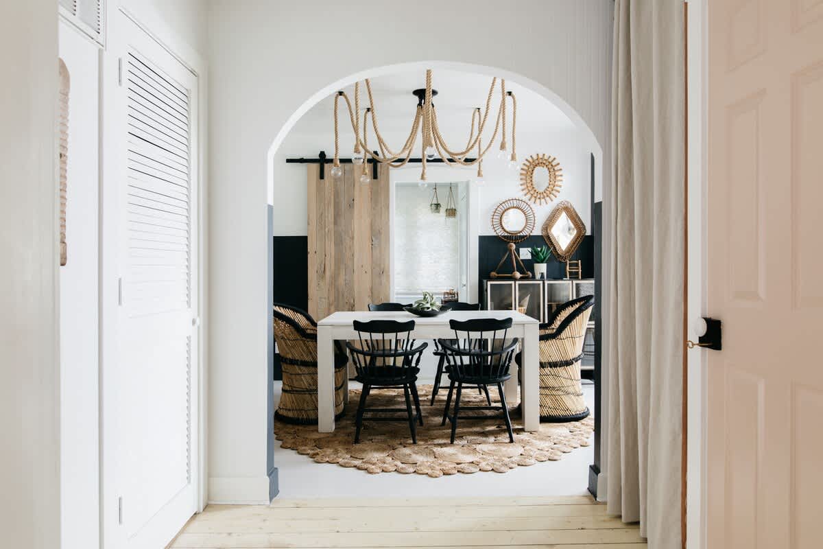 A dining room viewed through an arched doorway, featuring a white rectangular table with black spindle chairs, surrounded by woven chairs, a jute rug, and a rope chandelier, with a sliding barn door in the background.