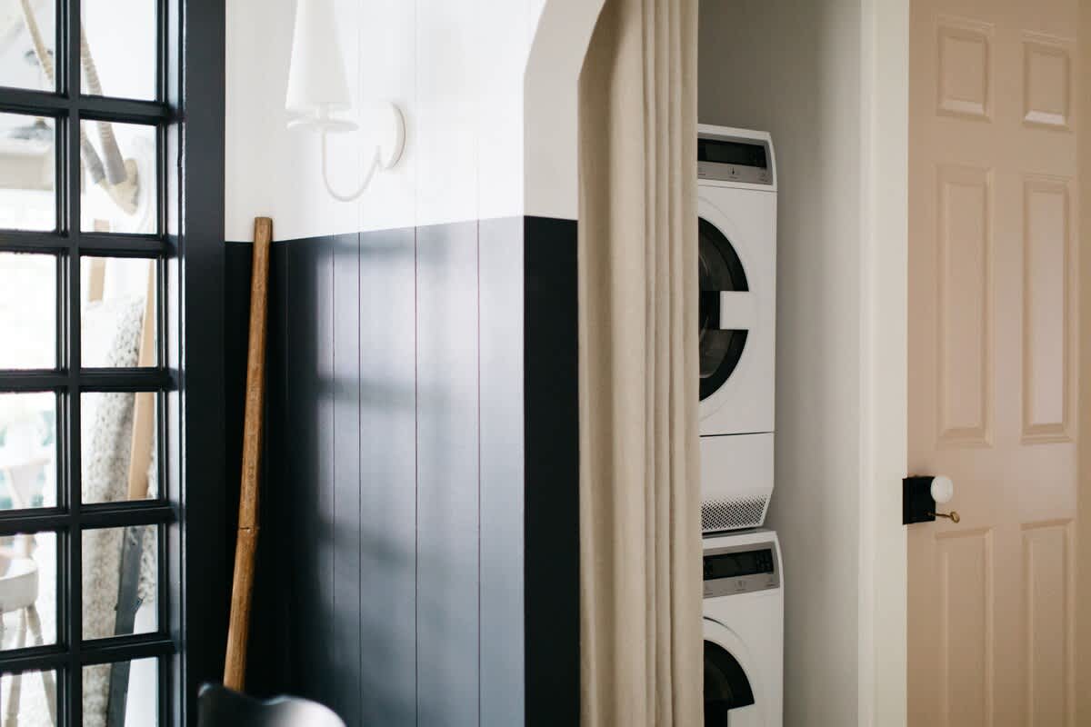 Compact laundry area with a stacked white washer and dryer, partially concealed by a beige curtain. The wall features modern black wainscoting contrasting with white upper walls, adjacent to a window with black grid panes.