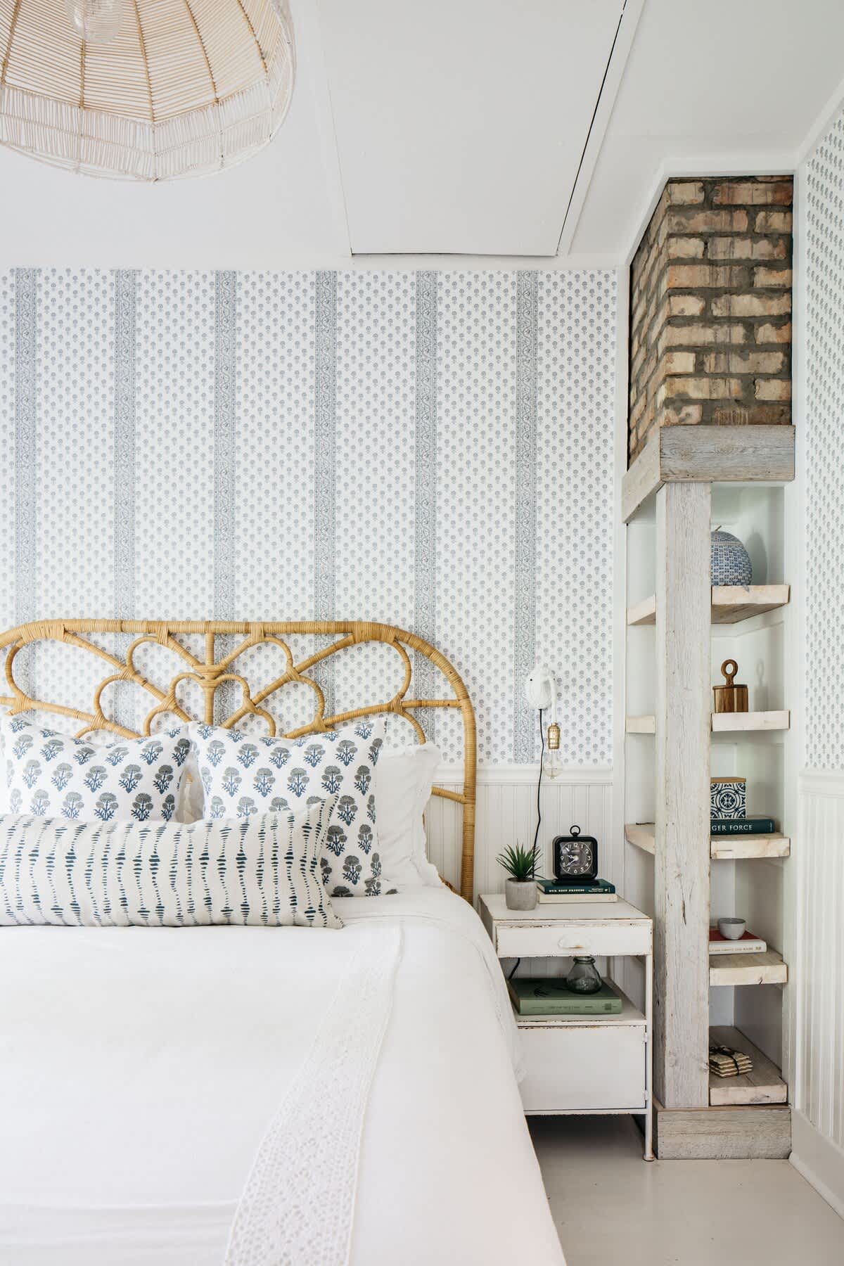 A cozy bedroom corner showcases a rattan headboard, patterned pillows, a small nightstand with a plant and alarm clock, and a built-in shelf next to an exposed brick chimney, adding rustic charm.