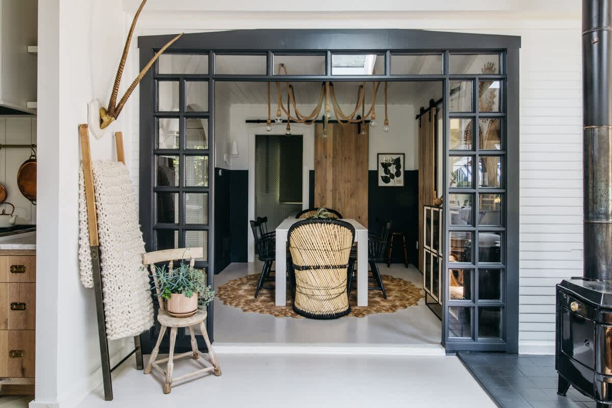 A modern dining room showcases a long white table, black and natural woven chairs, a unique rope chandelier, and large black-framed glass doors that open to another living space.