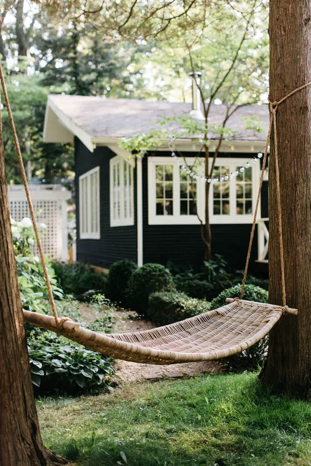 Relaxing woven hammock swing suspended between two trees in a lush green yard, with the dark-sided cottage visible in the background.
