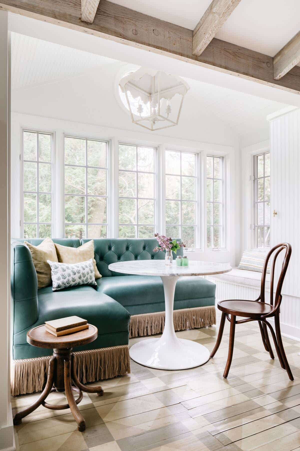 A cozy dining nook features a teal velvet L-shaped banquette with fringe, a white tulip table, and a single wooden chair, surrounded by large windows that offer bright natural light and views of greenery.