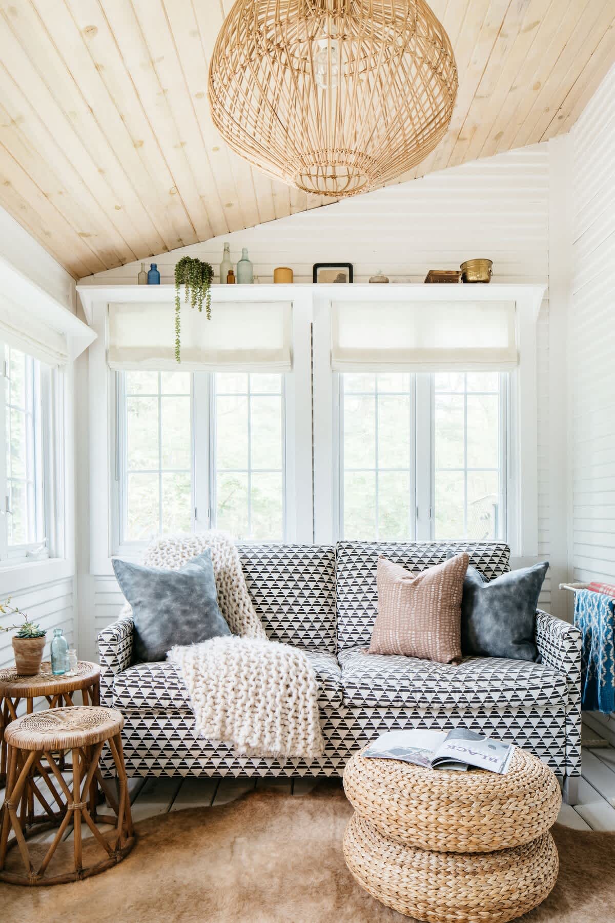 Cozy sunroom with a patterned sofa adorned with pillows and a chunky knit throw, a large woven light fixture, and multiple windows with white blinds. A small rattan side table and woven ottoman complete the space.