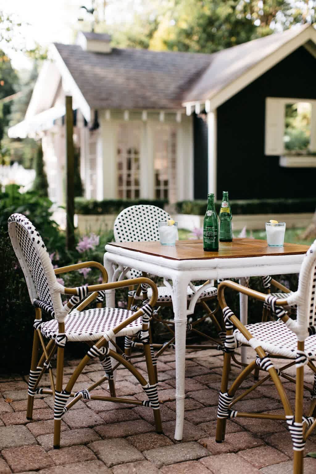 An inviting outdoor dining area on a brick patio features a white table with a wooden top and stylish black and white woven chairs, set against a backdrop of a dark house and lush garden foliage.