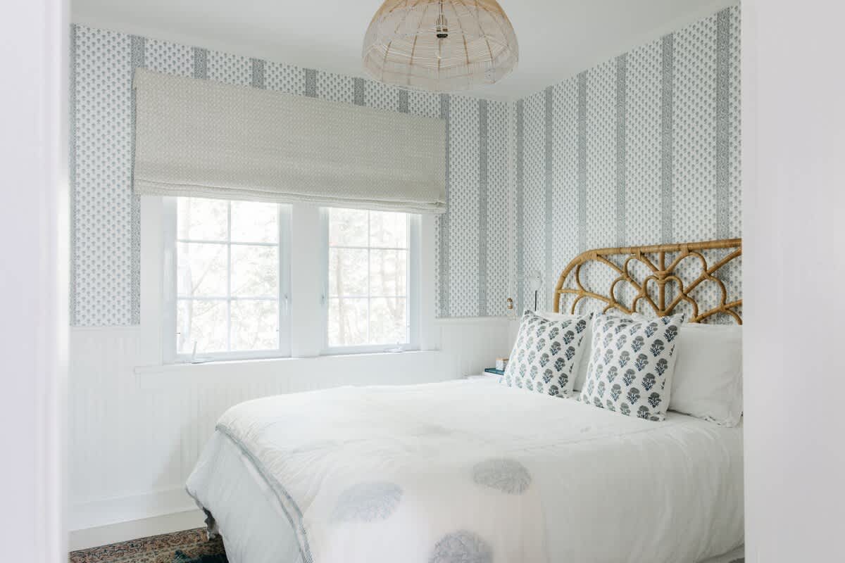 A serene bedroom features light blue and white striped wallpaper, a decorative rattan headboard, crisp white bedding, and two windows with a Roman shade, creating a tranquil and airy space.
