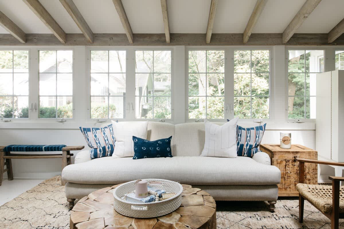 A bright living room with a vaulted ceiling and exposed wooden beams, featuring a cream striped sofa with blue and white pillows, a large tree-stump coffee table, and large windows overlooking greenery.