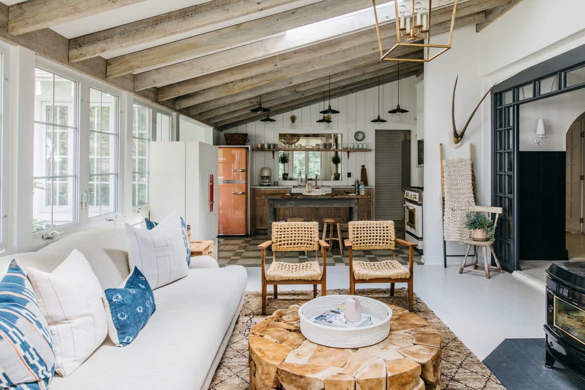 An open-concept living space with a vaulted ceiling and exposed wooden beams, featuring a cream sofa with blue and white pillows, a large tree-stump coffee table, and two woven armchairs, with a kitchen visible in the background.
