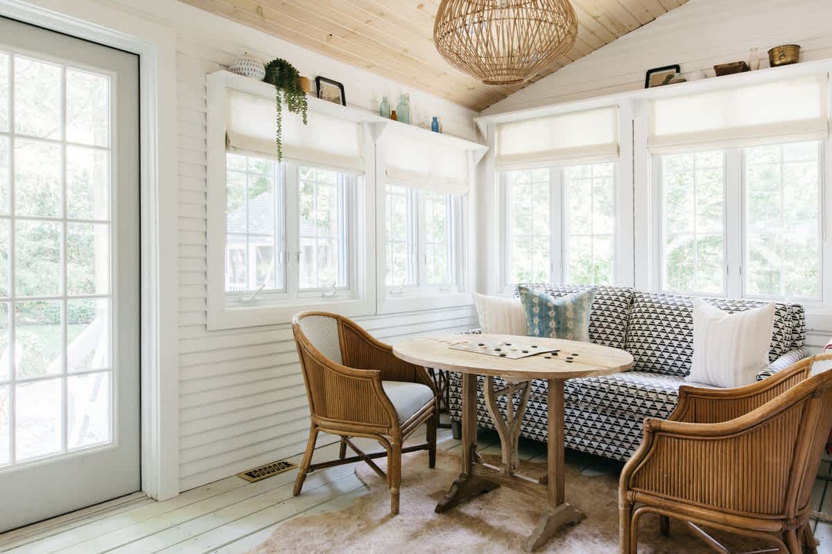 Bright sunroom with a patterned sofa, a round wooden table with two rattan chairs, and multiple windows with white blinds. A large woven light fixture hangs from the slatted wood ceiling, and a glass door leads outside.
