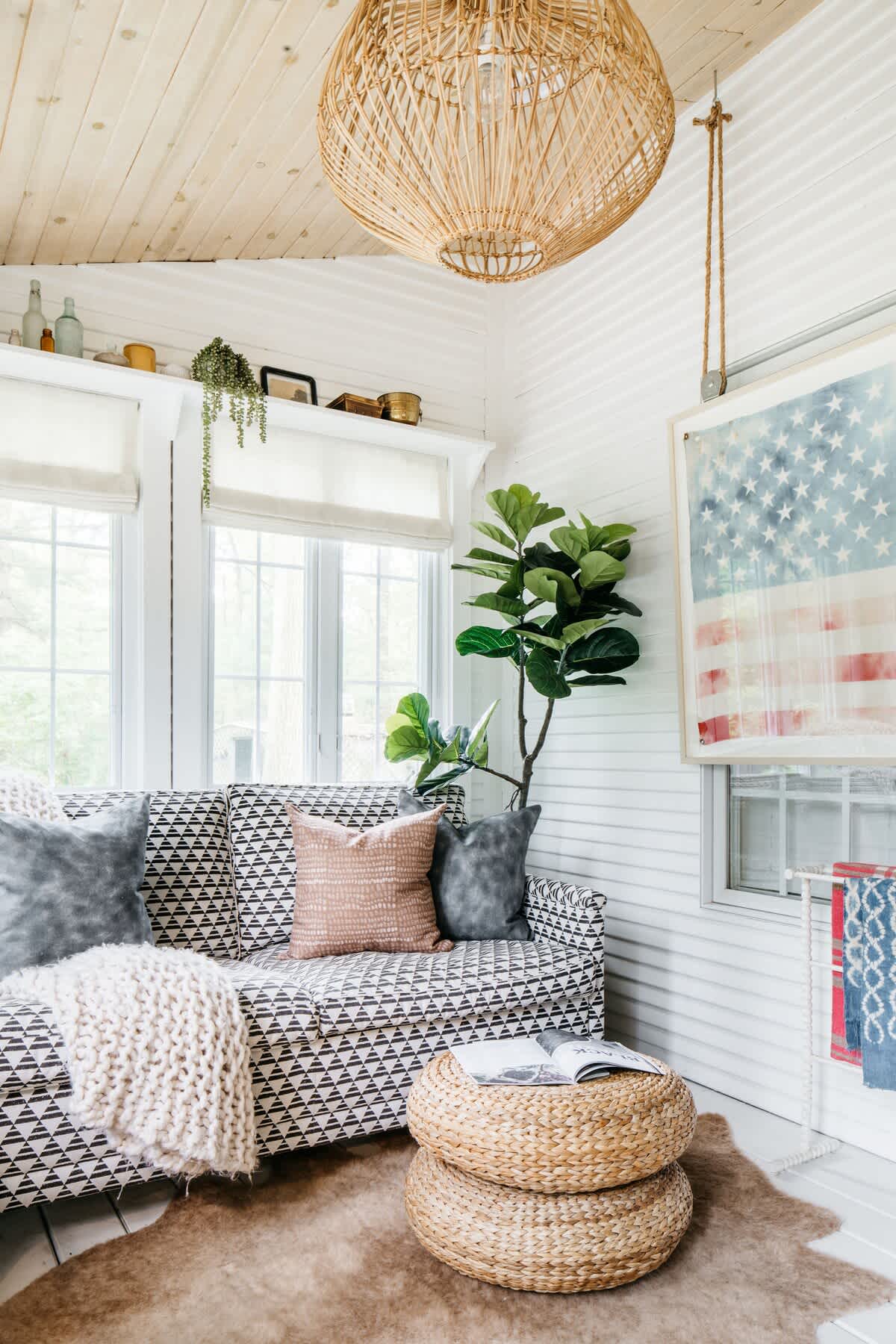 A bright sunroom with white paneled walls and ceiling, featuring a patterned sofa with throw pillows, a stacked woven ottoman, a large fiddle leaf fig plant, and a woven pendant light, with an American flag art piece on the wall.