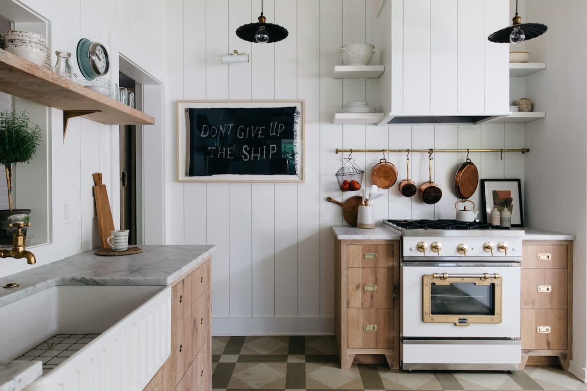 A charming kitchen with white paneled walls, checkered floor tiles, and light wood cabinetry with marble countertops. A white and brass stove is prominent, alongside a farmhouse sink and a "Don't Give Up The Ship" sign.