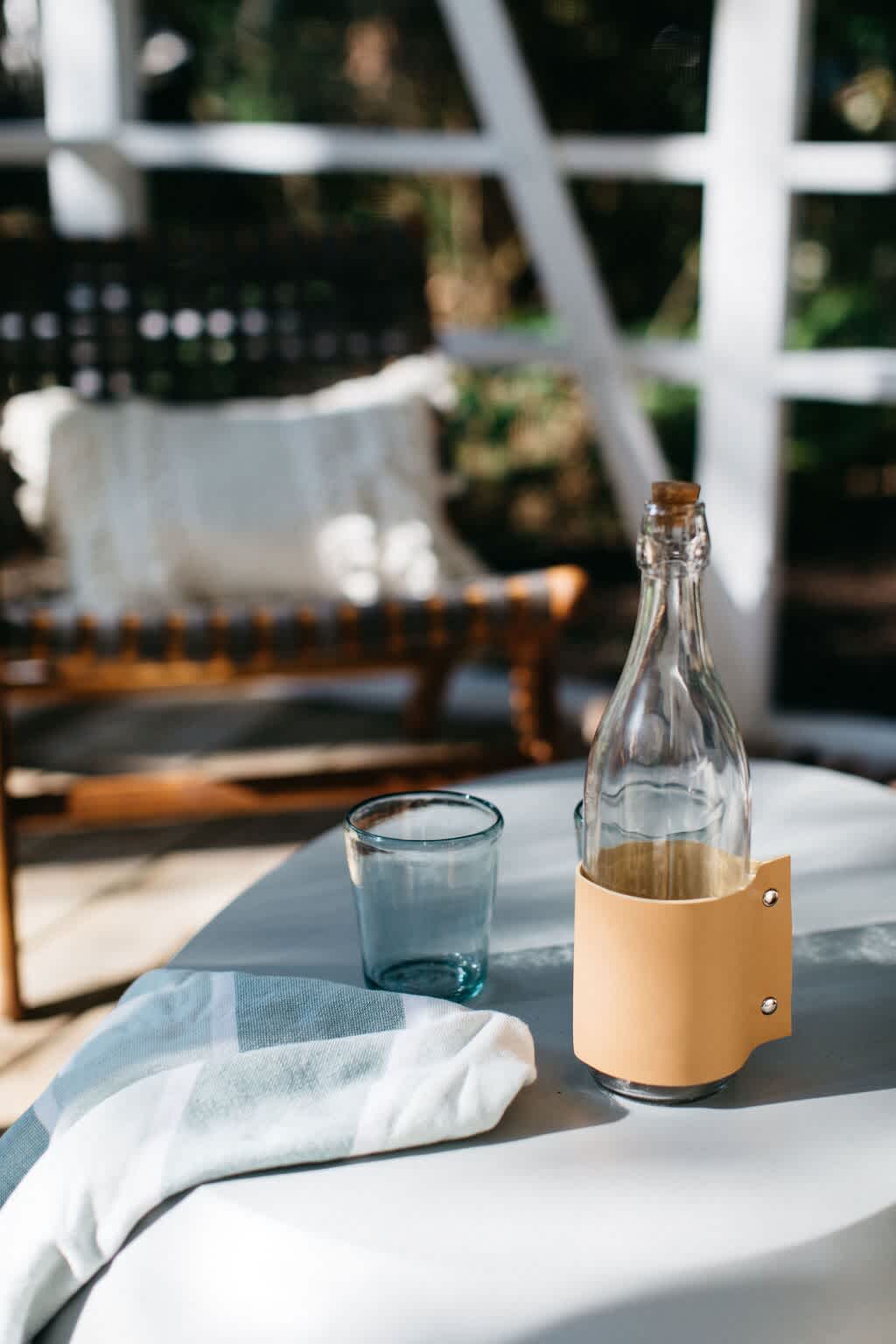 A close-up shot of a glass water bottle with a leather holder and a blue drinking glass on a light-colored table. A folded patterned towel is also visible, with blurred outdoor seating in the background.