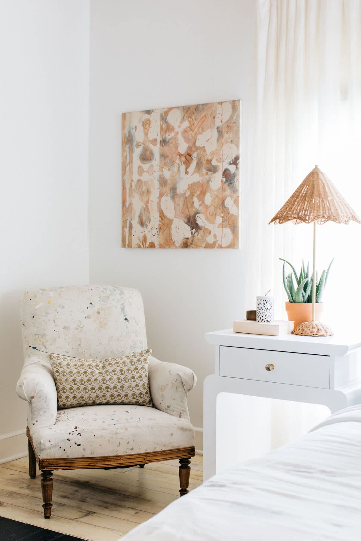 A close-up of a cozy corner in a bedroom, featuring a vintage upholstered armchair with a patterned pillow, a white nightstand with a wicker lamp and a potted plant, and abstract art on the wall.