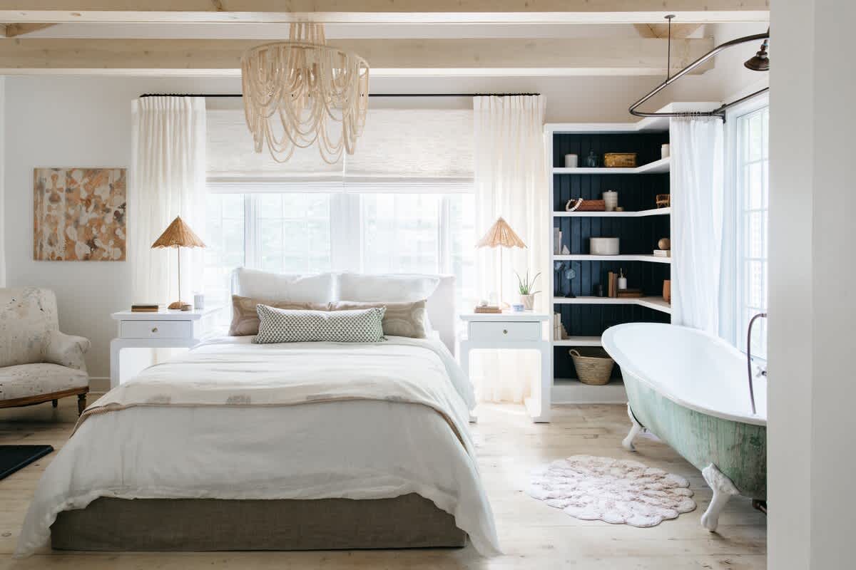 A spacious bedroom with light wood floors and a large window, featuring a white bed with a neutral headboard, two white nightstands with wicker lamps, a large woven chandelier, and a vintage-style bathtub in the corner.