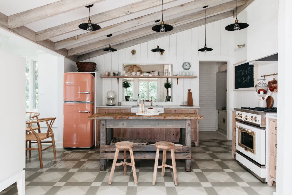A rustic-chic kitchen boasts a prominent peach-colored refrigerator, a large wooden island with two stools, white shiplap walls, exposed wooden ceiling beams, and light-toned patterned floor tiles.