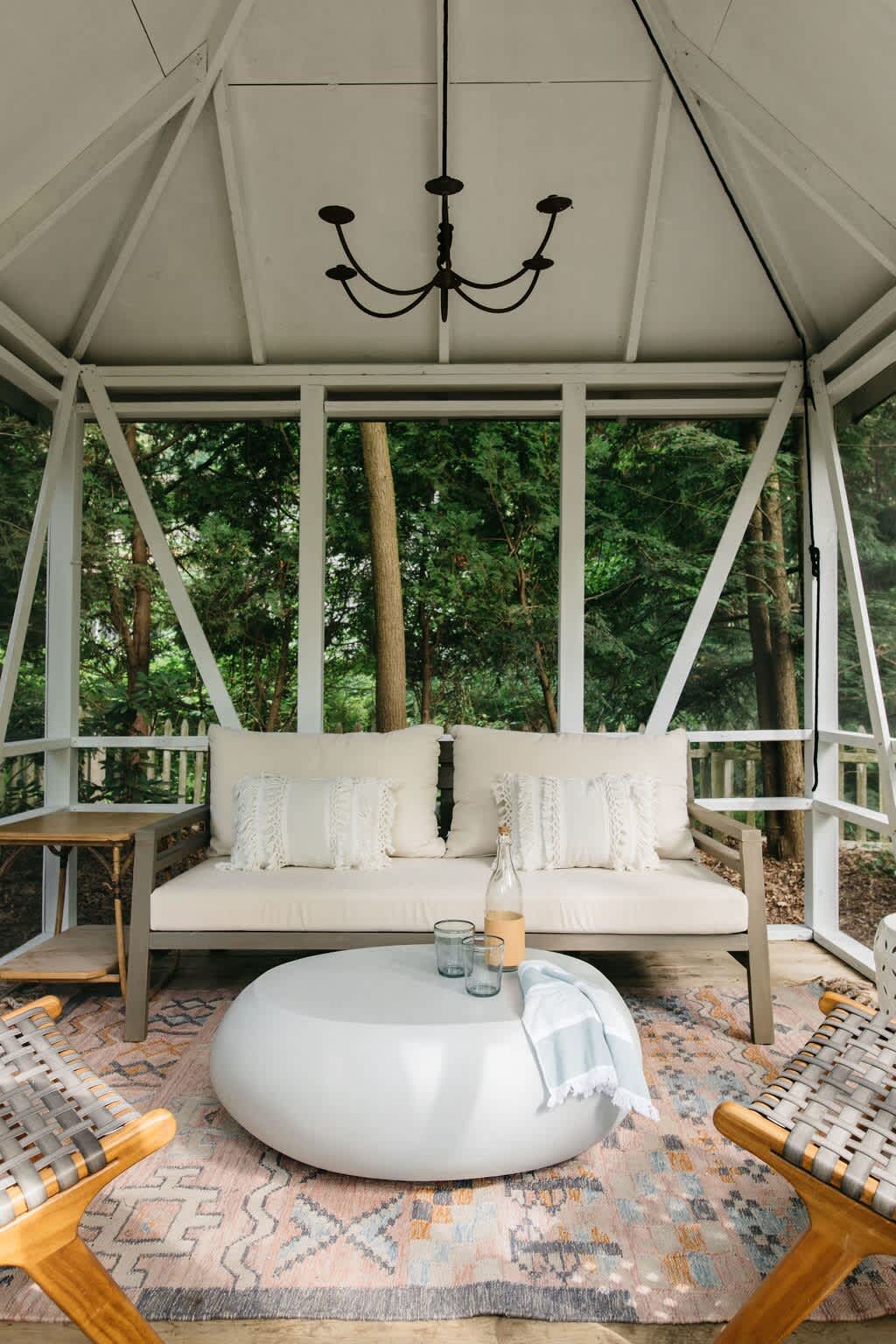 A cozy outdoor gazebo features a white slatted ceiling with a dark chandelier, a light-colored outdoor sofa with cream pillows, a round white coffee table, and a patterned rug, surrounded by lush green trees.
