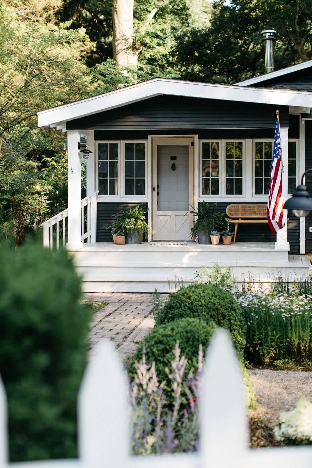 The front exterior of a dark-colored cottage with white trim, a welcoming porch with a white railing, a light-colored front door, and an American flag, surrounded by lush greenery and a picket fence.