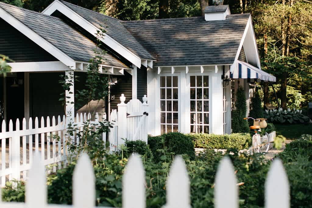 Charming cottage exterior with dark siding, white trim, and a white picket fence, surrounded by lush green landscaping and a vintage bicycle parked nearby.