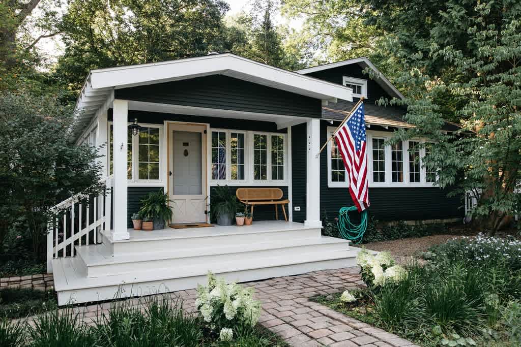 Charming dark green cottage with white trim, a welcoming front porch, and an American flag. The porch features a light wood bench, potted plants, and white railings, surrounded by lush greenery and a brick pathway.
