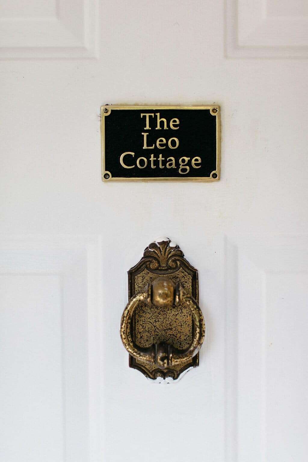 A close-up of a white door with a brass door knocker and a black and gold plaque that reads "The Leo Cottage".