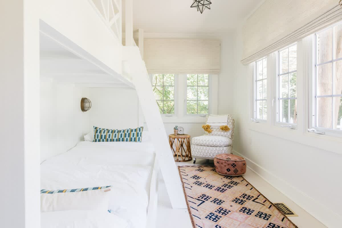 Bright, all-white bedroom featuring a built-in bunk bed with a ladder, a patterned rug, a cozy armchair, and multiple windows with natural light. A small wooden side table and a patterned ottoman add texture.