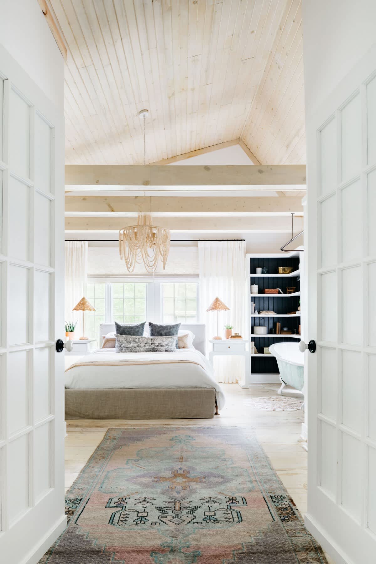 A spacious master bedroom features a large bed with neutral bedding, a light wood vaulted ceiling with exposed beams, a beaded chandelier, and a built-in black bookshelf next to a freestanding bathtub.