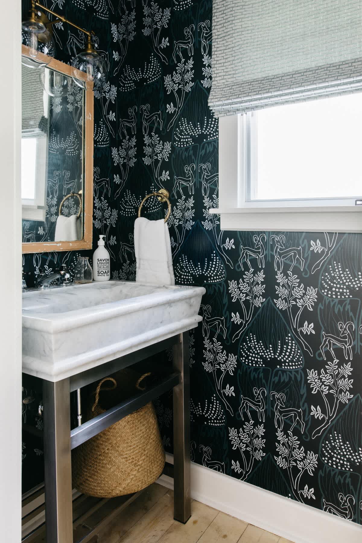 A stylish powder room features dark patterned wallpaper with deer and foliage, a marble-top vanity with a sleek metal base, a wooden-framed mirror, and a small window for natural light.