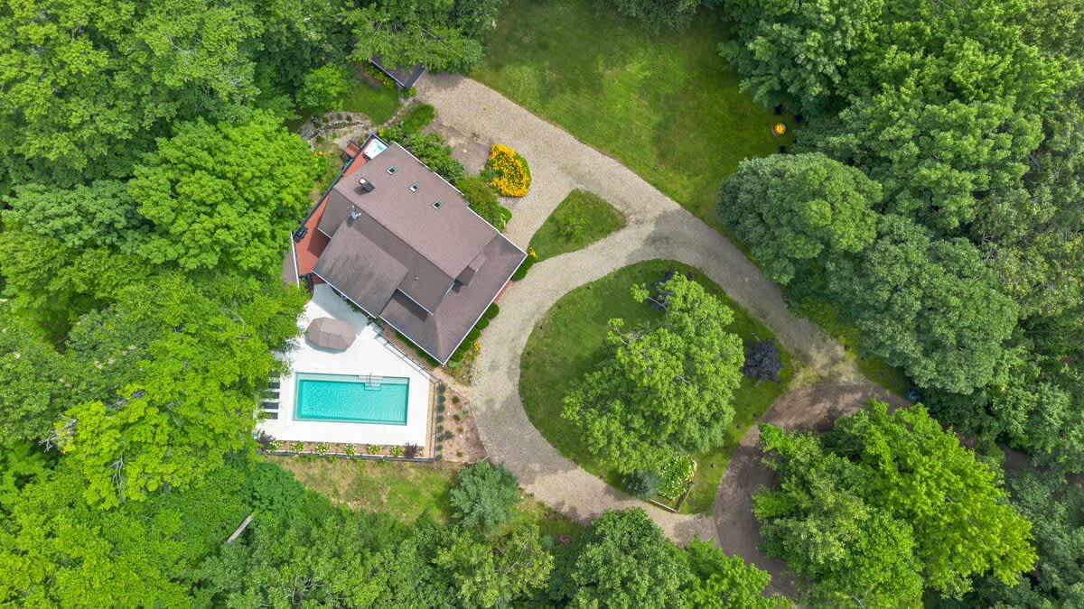 A high aerial view reveals the entire property, including a dark gray house, a winding gravel driveway, an outdoor pool, and a hot tub area, all completely enveloped by a dense, verdant forest.