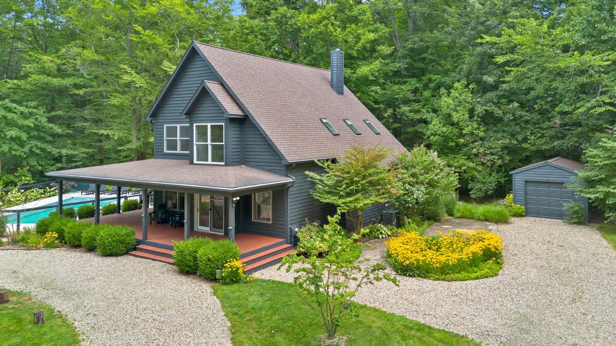 An aerial view showcases a dark gray two-story house with a brown roof, a gravel driveway, and an outdoor swimming pool area, all nestled within a lush green forest.