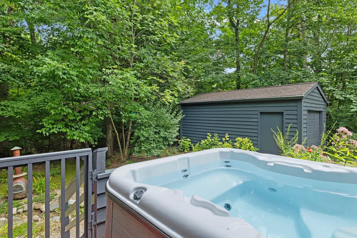 An outdoor hot tub with a dark wood surround is situated on a deck, surrounded by lush green trees and a dark gray shed in the background.