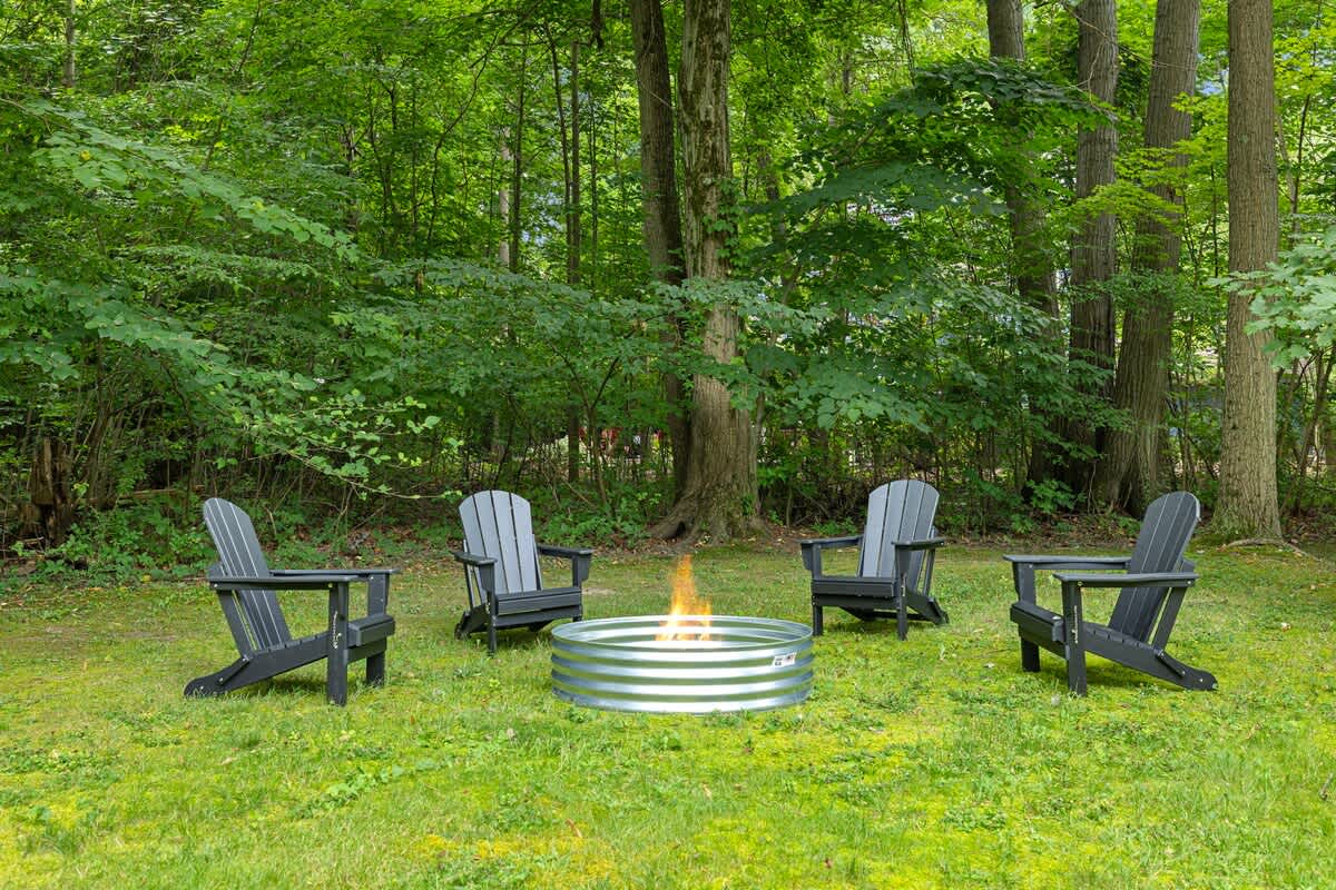 A circular metal fire pit with a burning fire is centered on a grassy lawn, surrounded by four dark gray Adirondack chairs, all nestled within a dense forest of tall green trees.