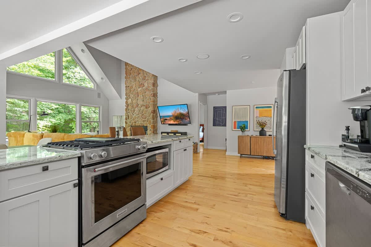 View from the kitchen, showcasing light gray cabinets, stainless steel appliances, and an open layout leading to the living room with a prominent stone fireplace and large windows.