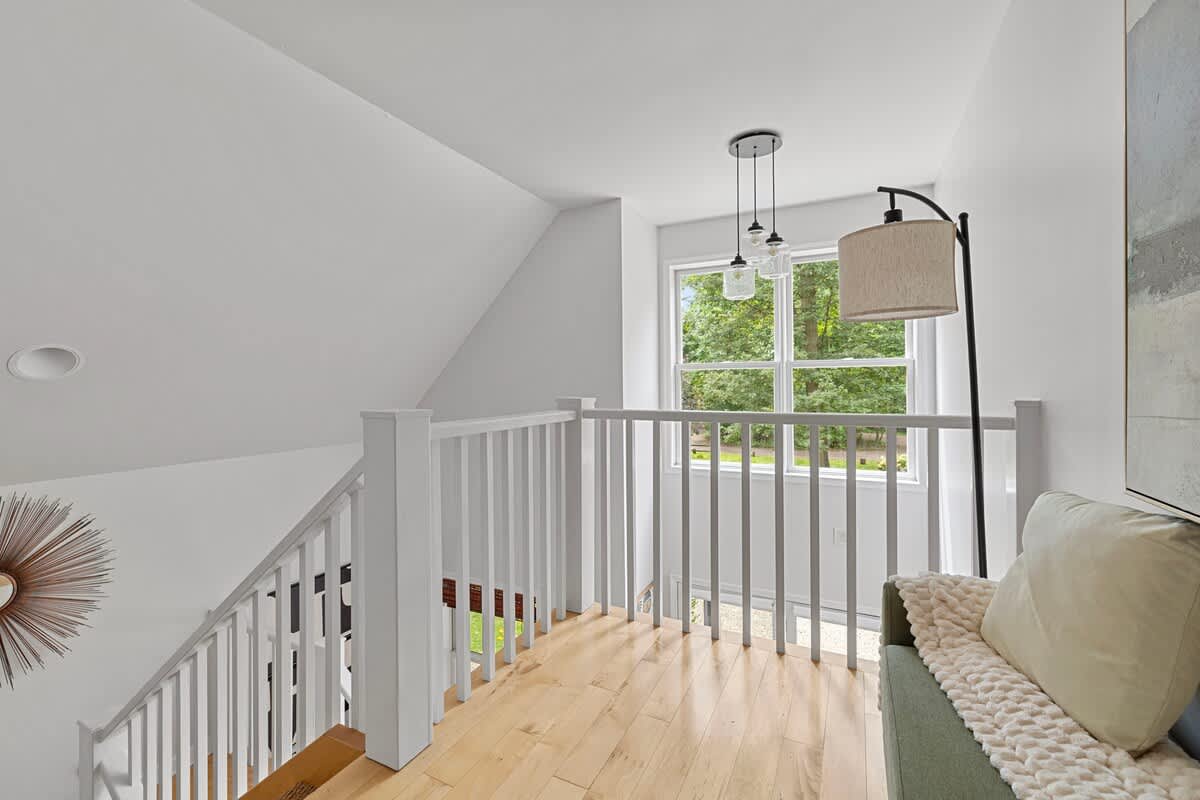 An airy upstairs landing with white balusters and light hardwood floors, featuring a cozy reading nook with a floor lamp and a large window offering views of the surrounding trees.