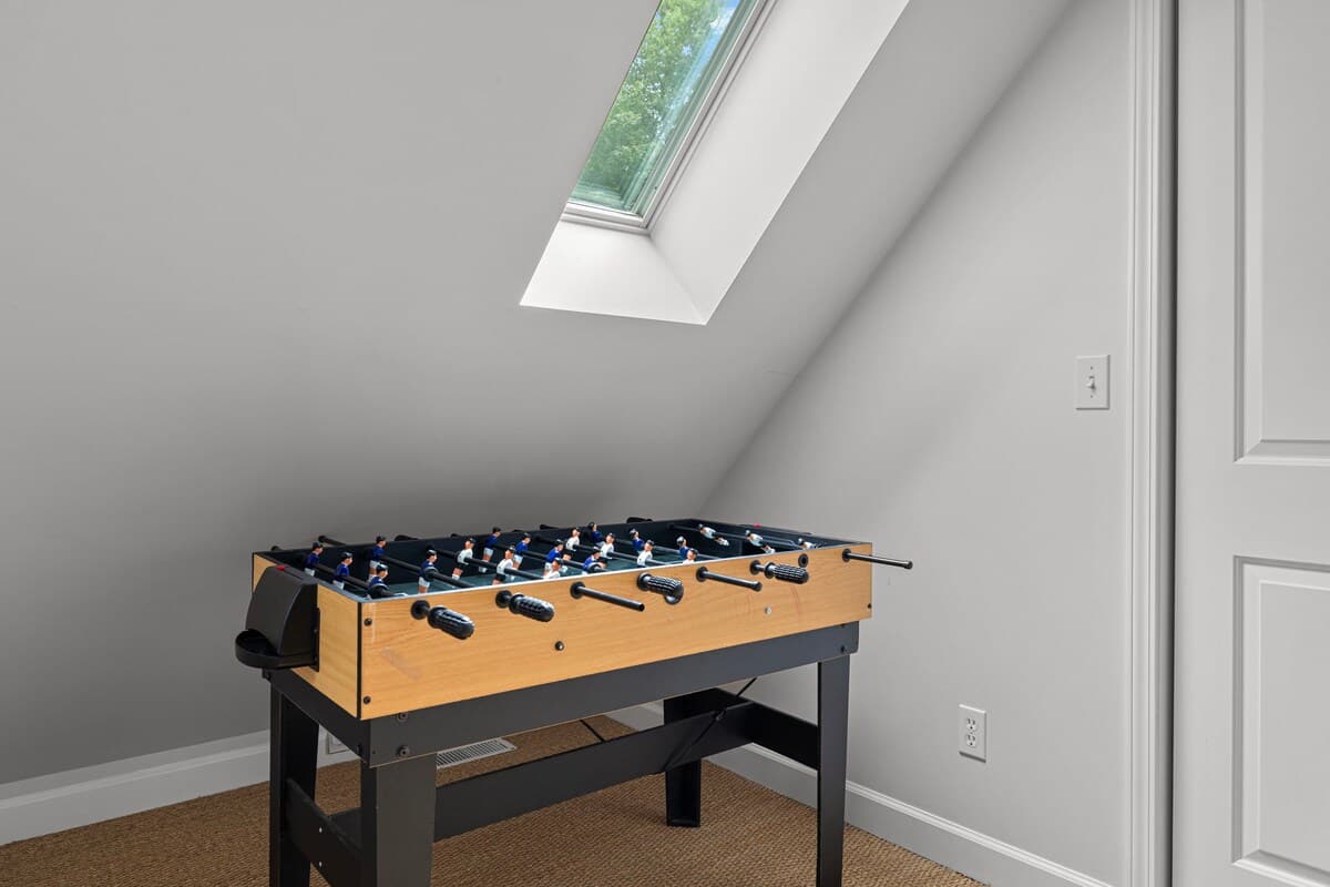 An attic game room with a foosball table, light gray walls, and a large skylight offering views of green trees. The floor is covered with a natural fiber rug.