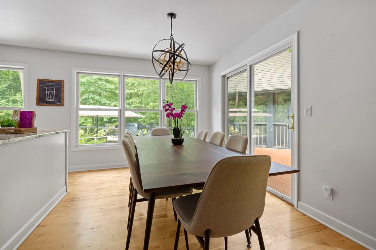 Bright dining area featuring a long dark wooden table, upholstered mid-century modern chairs, and a unique spherical chandelier, surrounded by large windows and a sliding glass door to the deck.