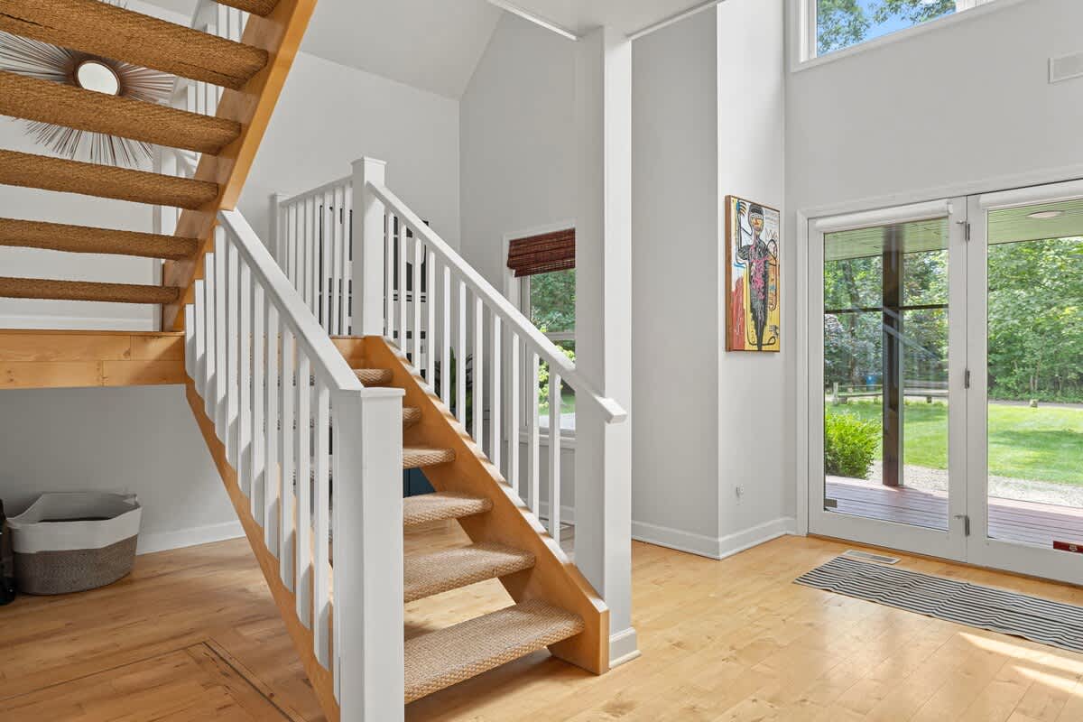 Spacious foyer featuring a light wood staircase with white railings and a woven runner, leading up to a second floor, with double glass doors providing access to the exterior.