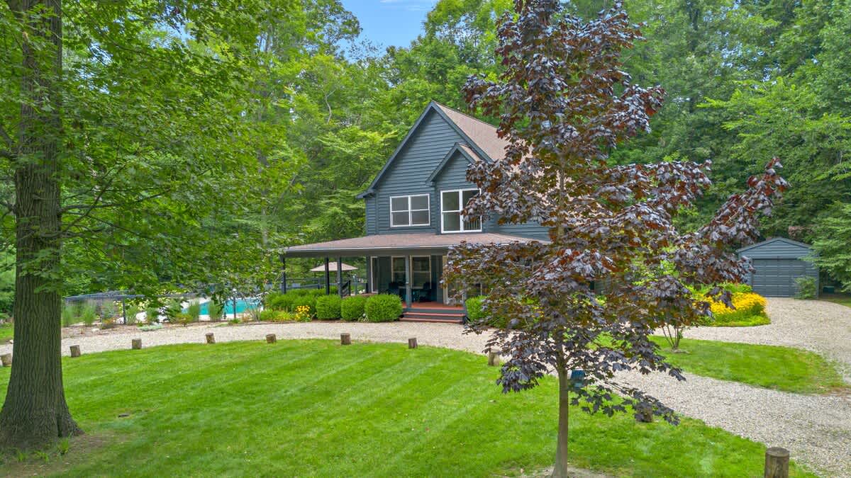 The front exterior of a dark gray two-story house with white trim and a covered porch, surrounded by a gravel driveway, manicured lawn, and mature green trees.