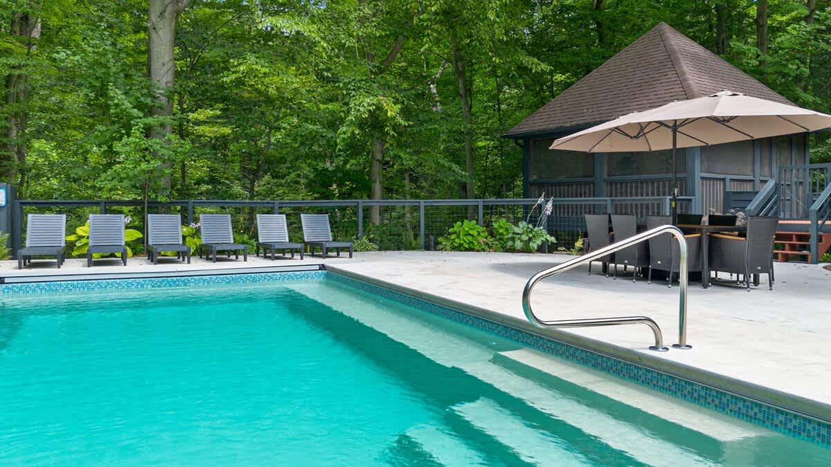 A sparkling blue outdoor swimming pool with a tiled edge is surrounded by a concrete patio, several lounge chairs, and a covered gazebo with dining furniture, all set against a backdrop of dense green trees.