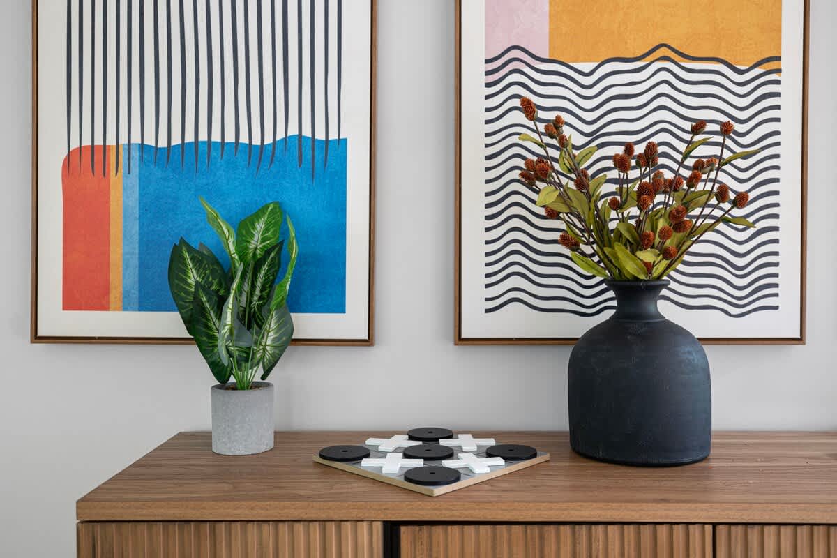 A wooden console table displays modern abstract art prints, a potted snake plant, a black decorative vase with dried flowers, and a stylish black and white tic-tac-toe board game.