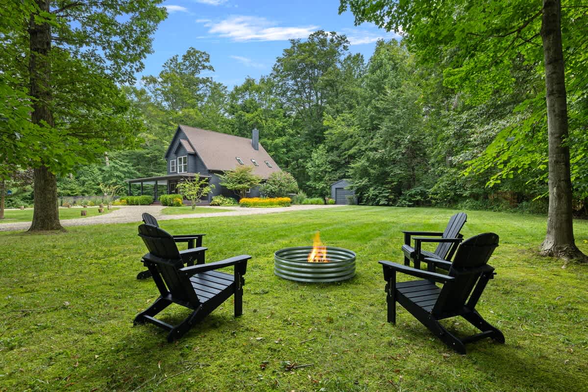 Outdoor fire pit area with a circular metal fire ring and four black Adirondack chairs on a grassy lawn, surrounded by mature trees with the house visible in the background.