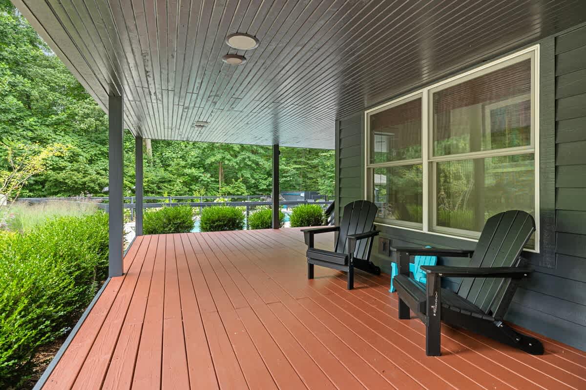 Covered outdoor porch with a dark wood deck, two black Adirondack chairs, and views of lush green bushes and the pool area beyond a railing.