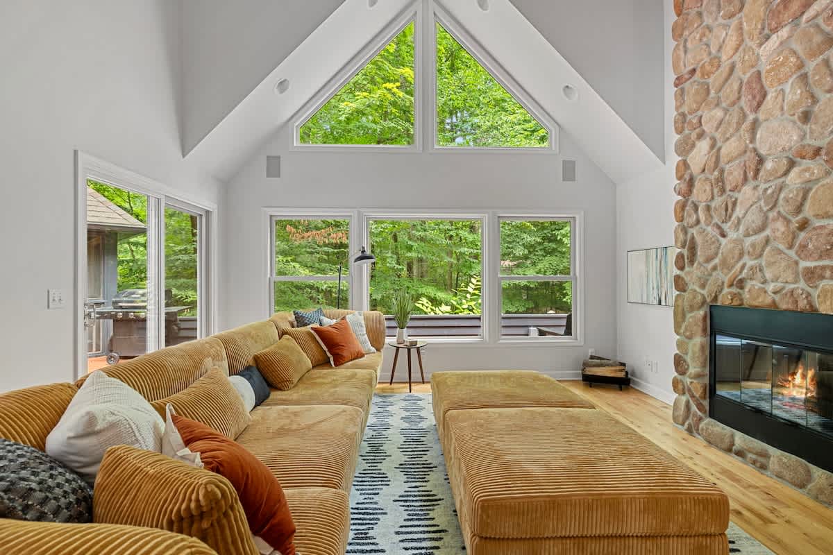 Spacious living room with a large corduroy sectional sofa, a prominent stone fireplace, and vaulted ceilings with large windows offering views of lush greenery.