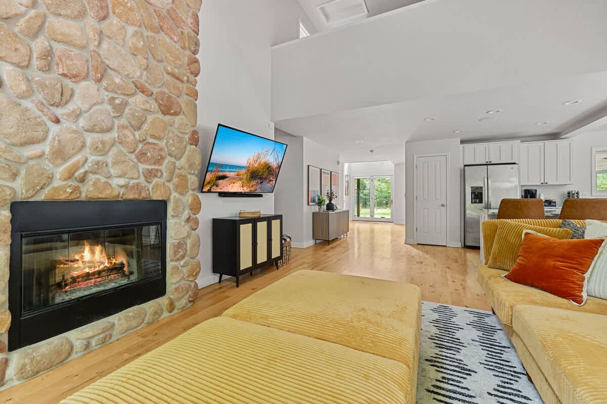 Inviting living room centered around a floor-to-ceiling natural stone fireplace with a black firebox, a wall-mounted flat-screen TV, and a comfortable mustard yellow sectional sofa with matching ottomans.
