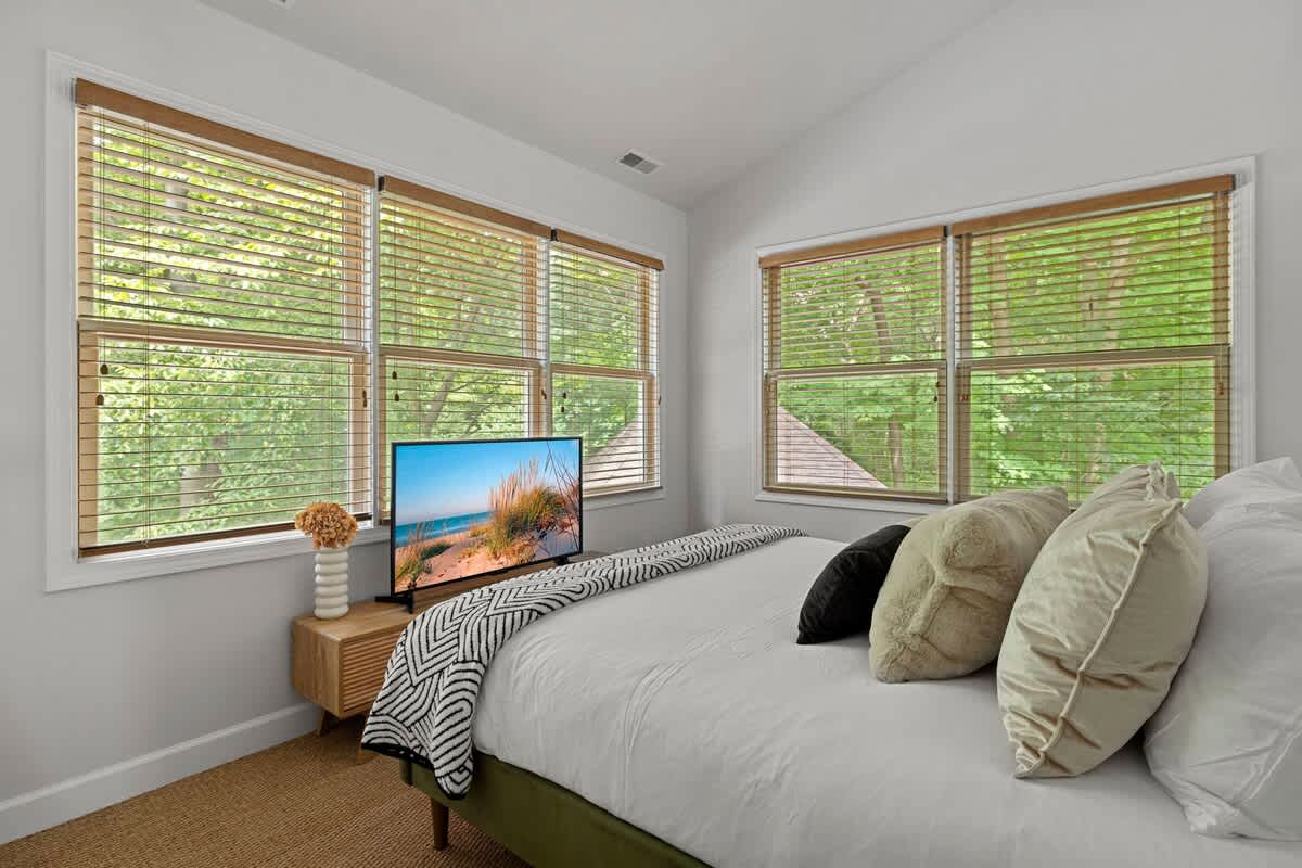 A cozy bedroom with a bed dressed in white and a patterned throw, facing a wall with multiple windows overlooking greenery. A flat-screen TV rests on a wooden console.