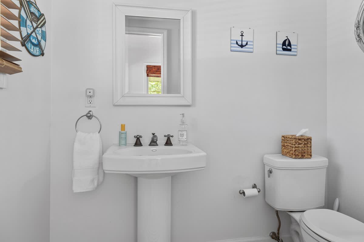 A bright white half bathroom features a pedestal sink with a white framed mirror above, nautical-themed wall decor, and a toilet with a woven tissue box.
