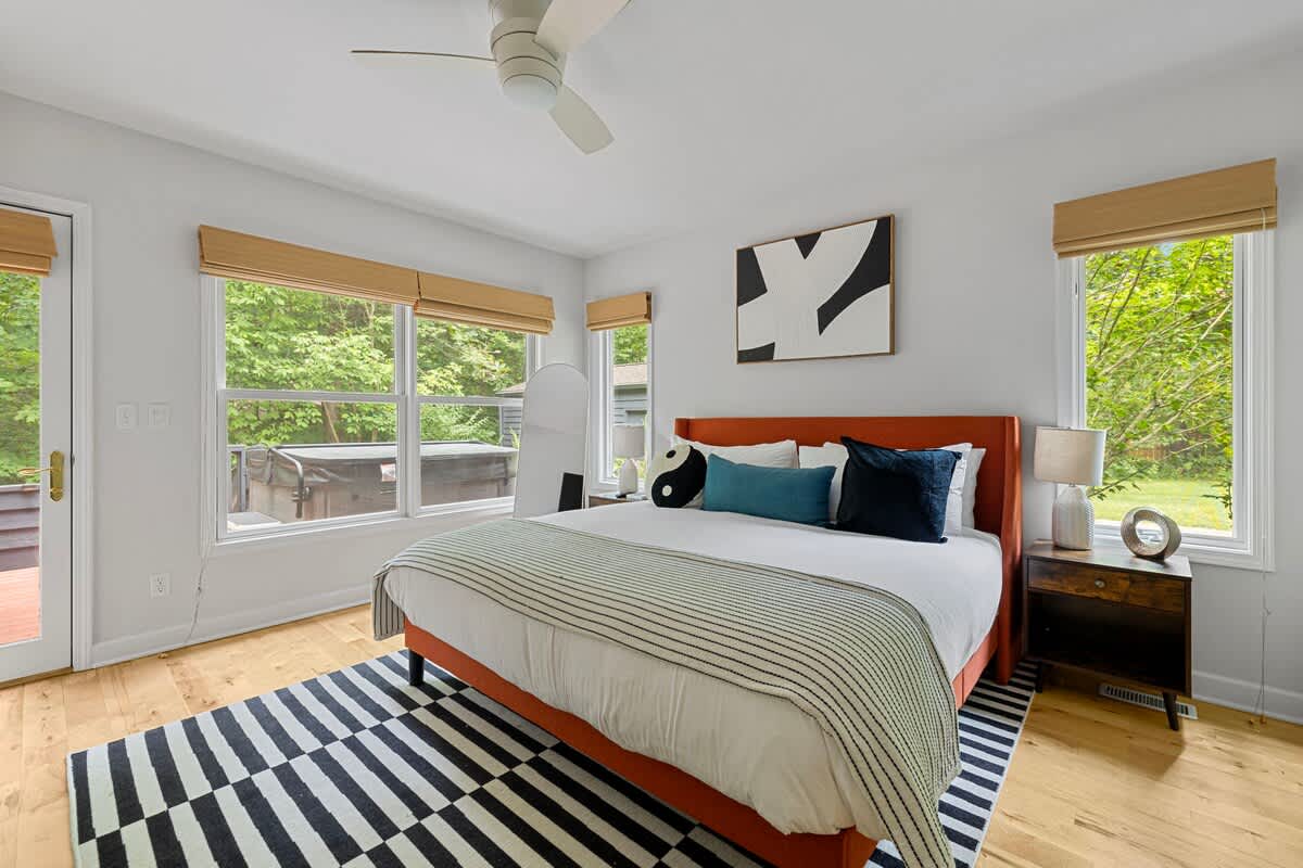 A spacious bedroom with an orange upholstered bed, a black and white striped rug, and large windows offering views of lush greenery. A ceiling fan is visible overhead.