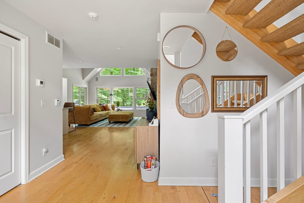 Bright entryway with light hardwood floors, a white staircase with a woven runner, and decorative mirrors on the wall, offering a glimpse into the living room.